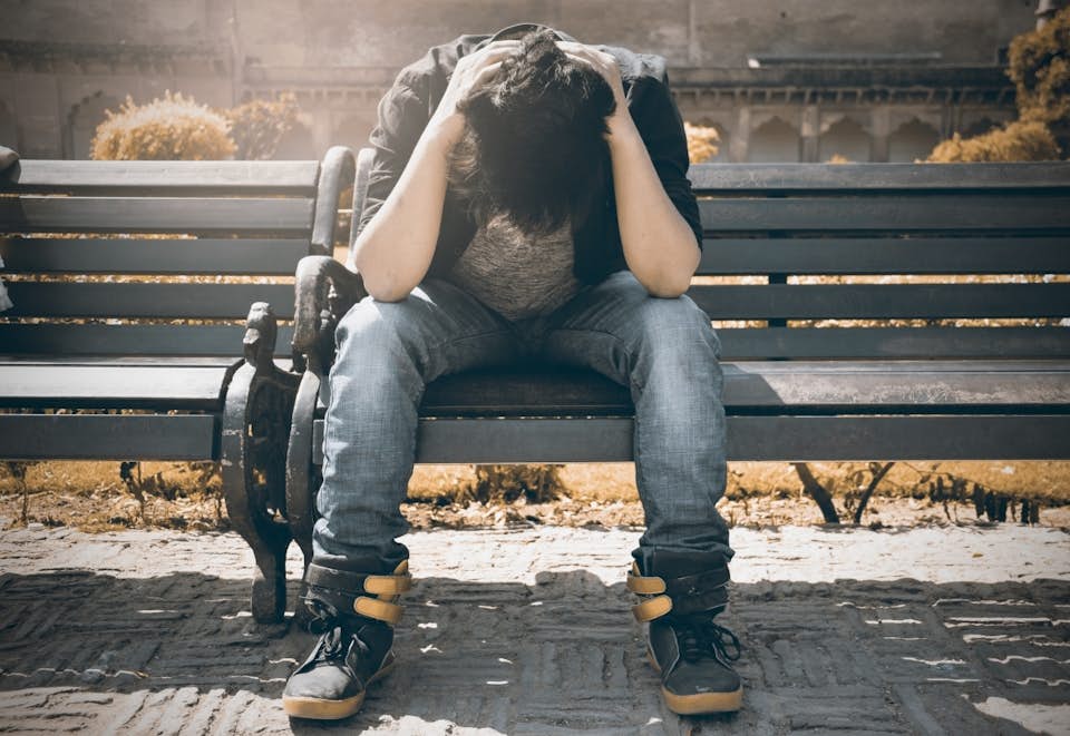 Depressed young man sitting on a park bench with his head in his hands.