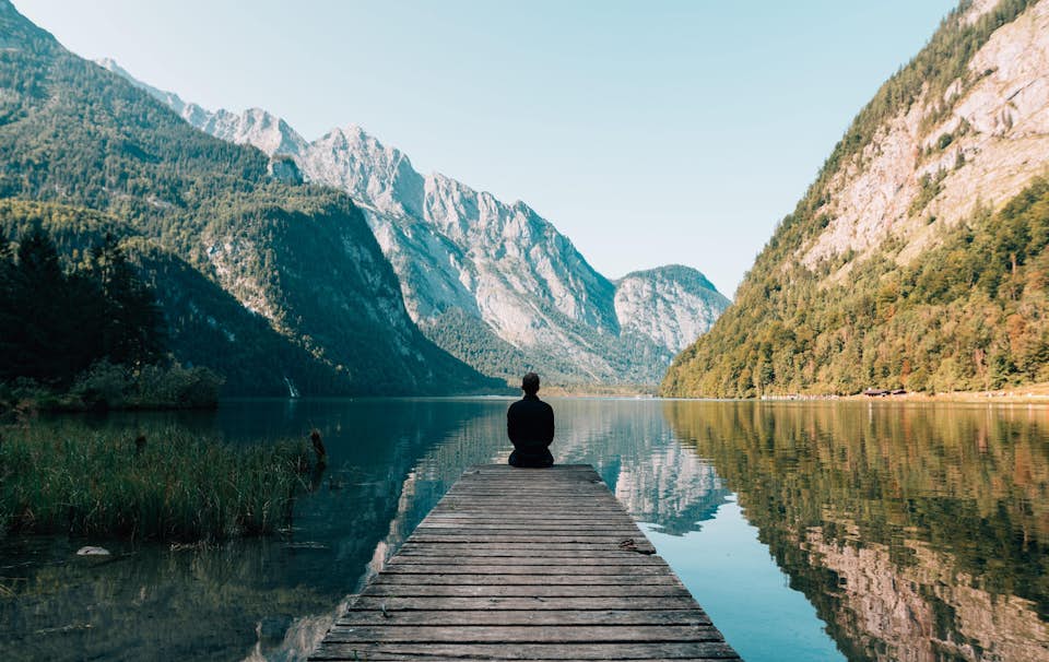 Relaxed man sitting on a dock overlooking mountains and a calm, still lake.